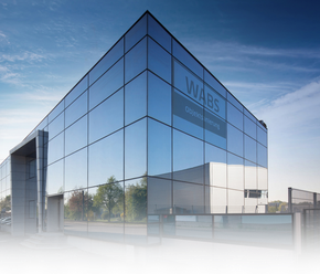 Modernes Glasgebäude mit „WABS“-Logo vor blauem Himmel, Bäume spiegeln sich in den Fenstern.