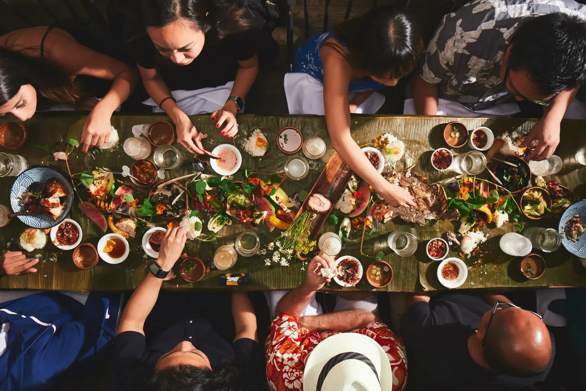 People gathered around a long, flower-filled banquet table, reaching for shared dishes and drinks from above.