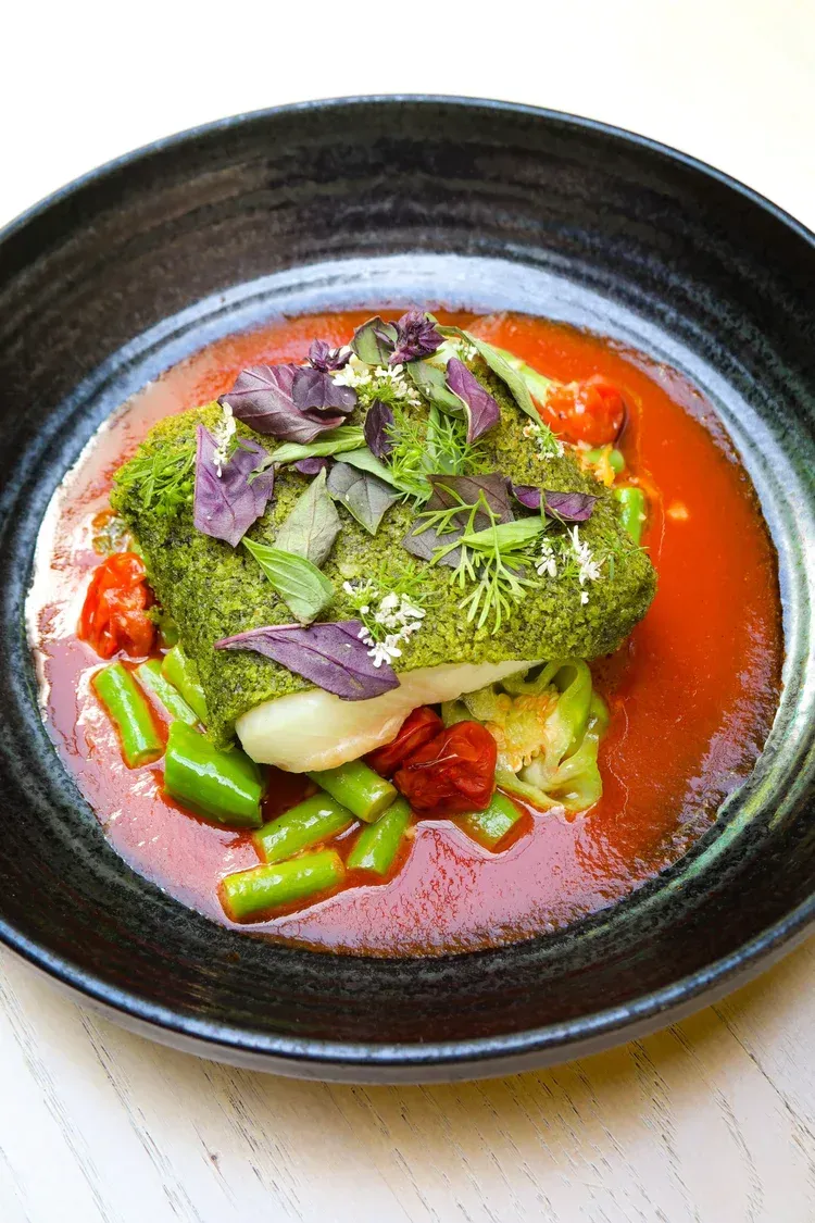 Gourmet dish with green herb-crusted entrée, vegetables, and red sauce in a black bowl