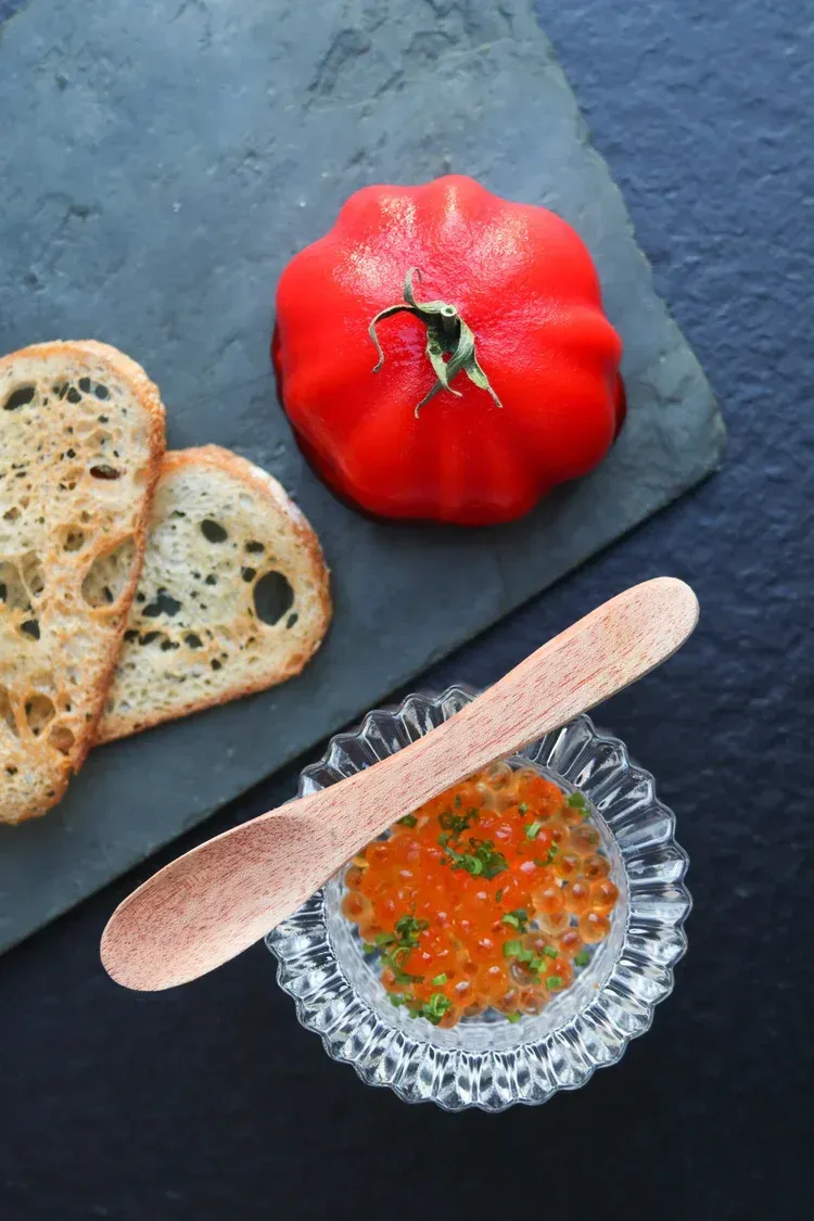 Tomato, sliced bread, and a bowl of red-orange caviar with a wooden spoon on a dark slate surface