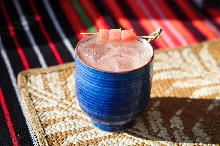 Blue cup with a pink drink and watermelon garnish on a woven mat