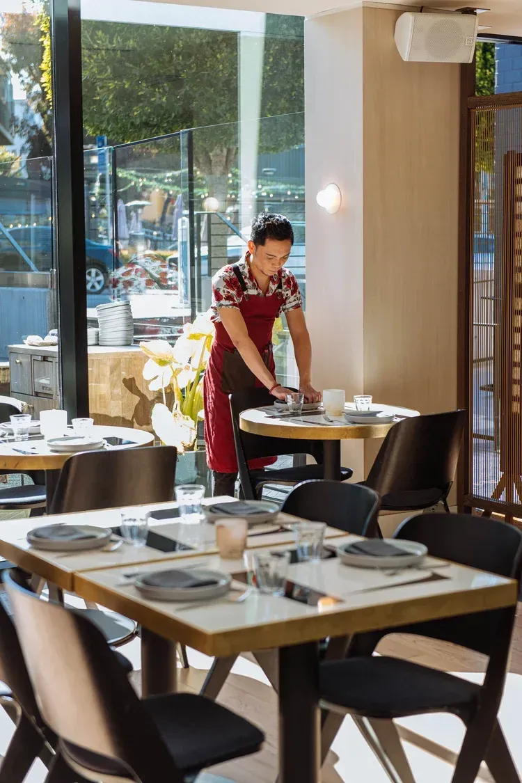 Waiter in a red apron setting a table in a bright restaurant with large windows.