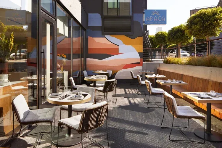 Outdoor café patio with tables, chairs, orange wall mural, and glass railings in sunlight