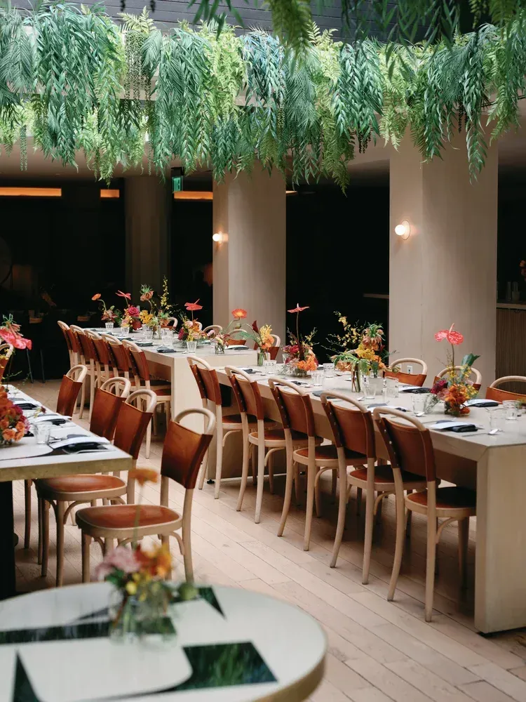 Long banquet table in a leafy outdoor dining space with wooden chairs and floral centerpieces