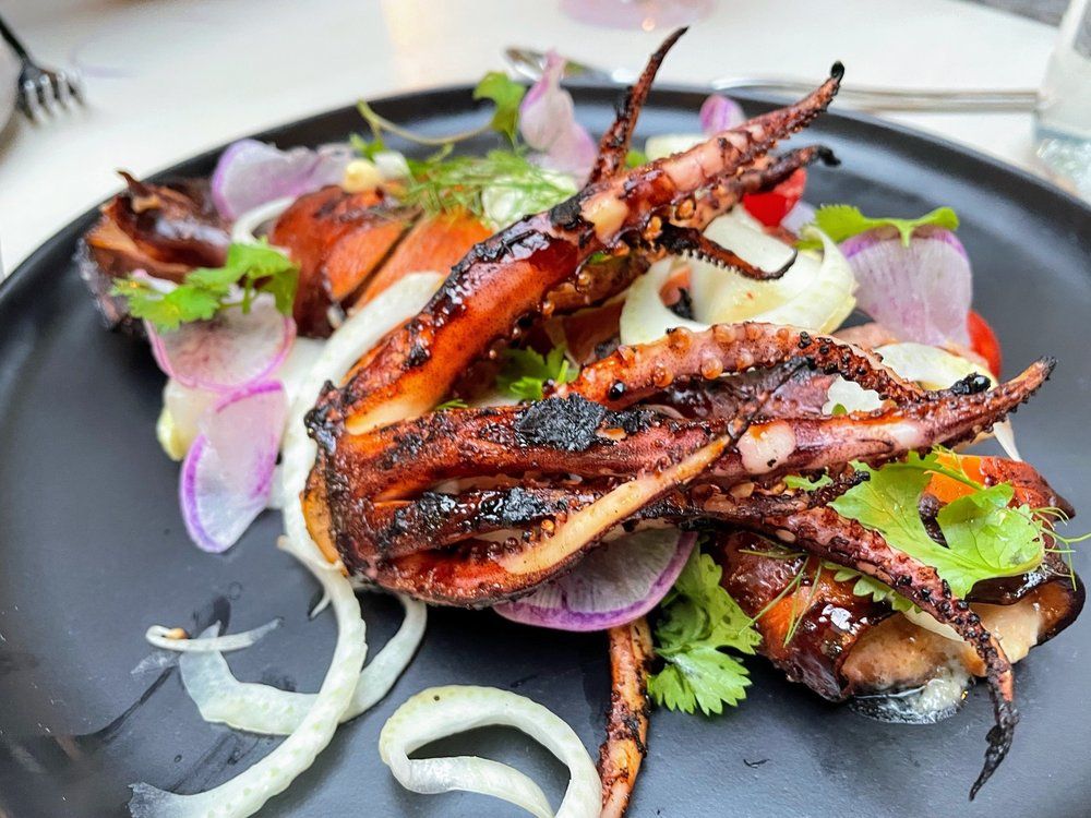 Grilled octopus tentacles on a dark plate with onion slices, herbs, and greens