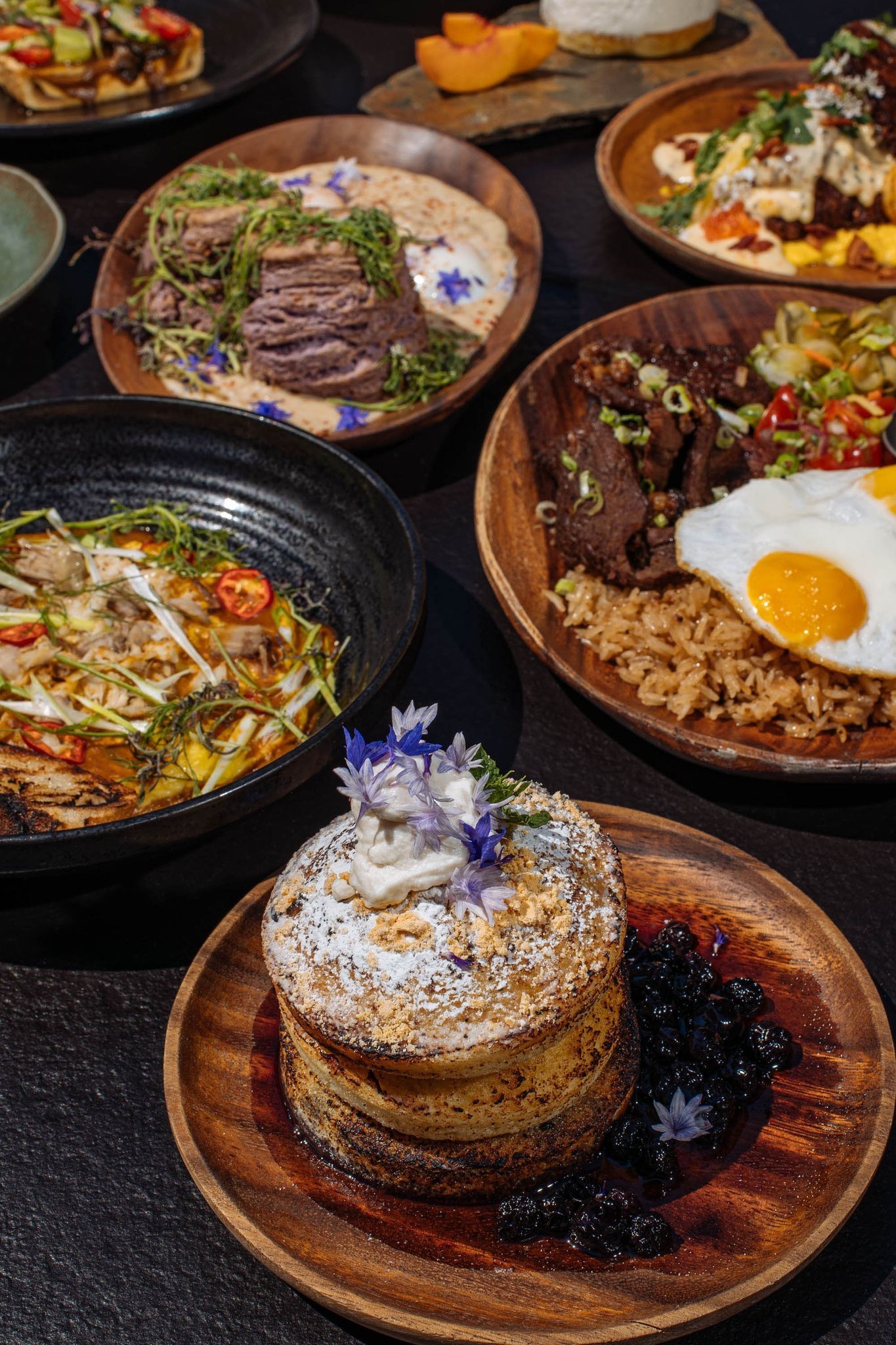 Assorted Japanese-style dishes on a dark table, including bowls with rice, noodles, beef, and a fried egg.