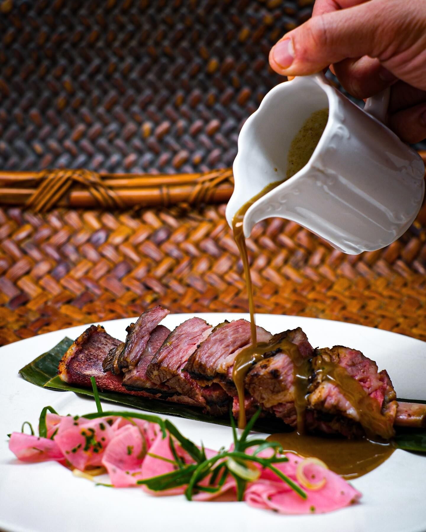 Sliced steak on a white plate with pink garnish, as sauce is poured from a small pitcher