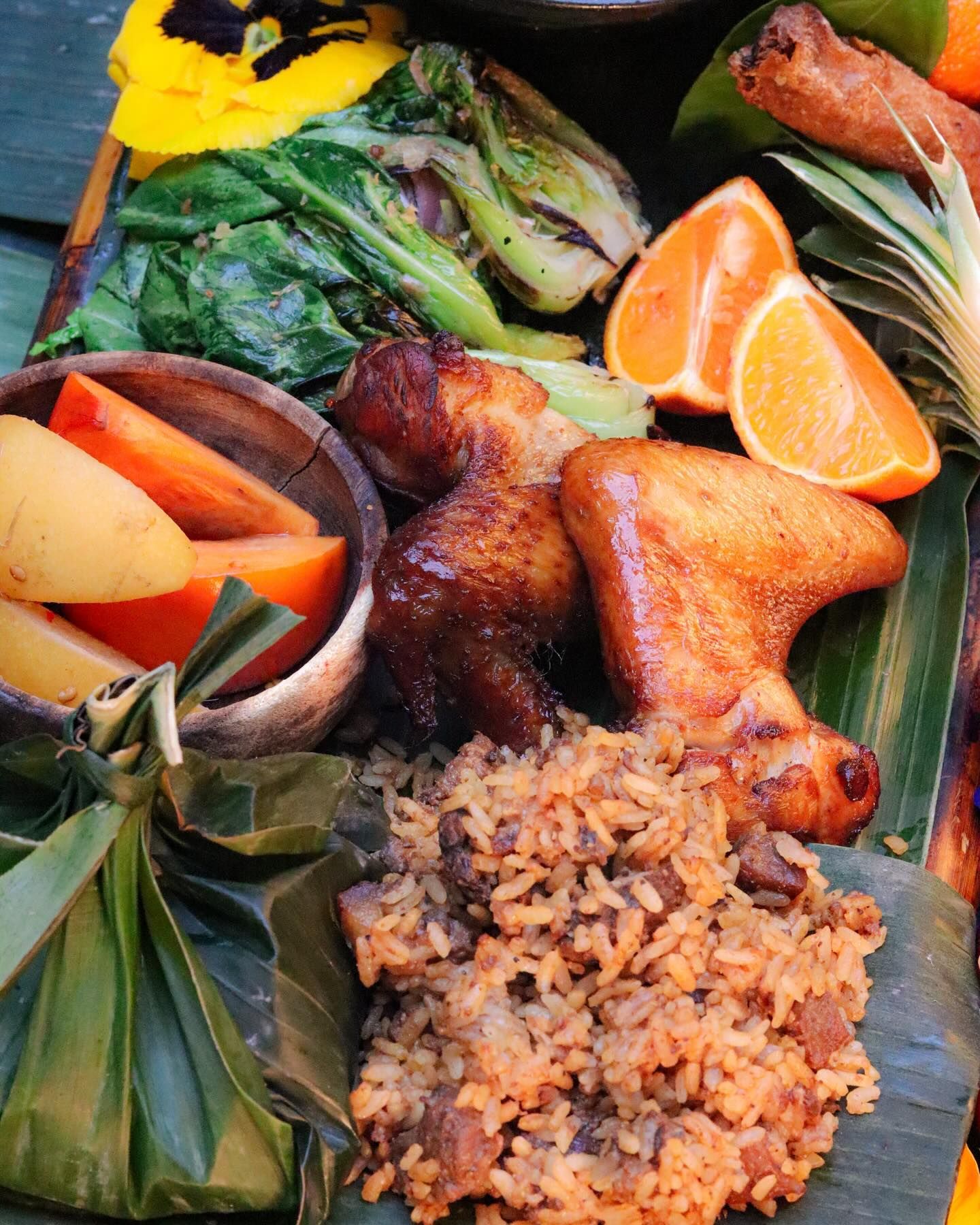 Assorted food platter with roasted chicken, rice, vegetables, and orange slices on banana leaves