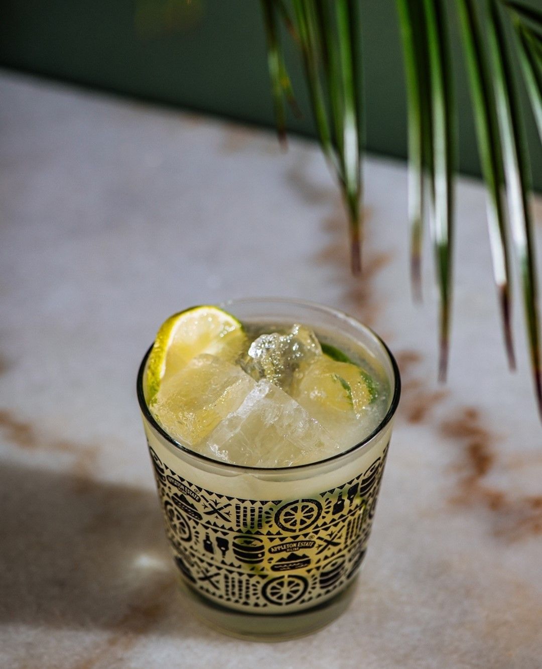 A patterned glass of a lemon cocktail with ice on a marble surface, with green plant leaves in the background