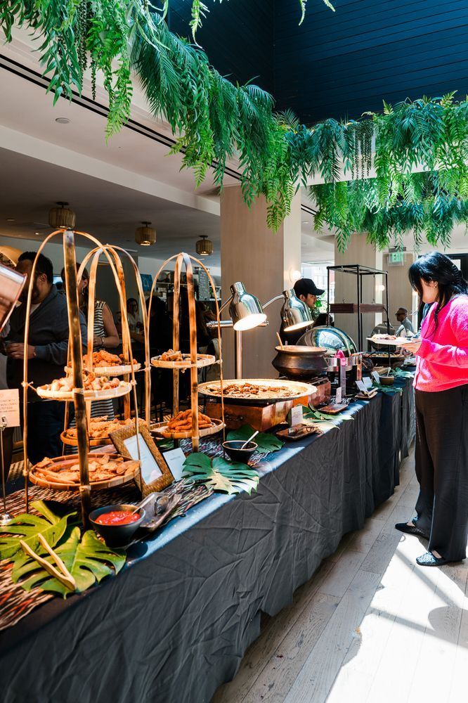 Outdoor buffet table under hanging greenery, with trays of food and a server in a bright pink jacket