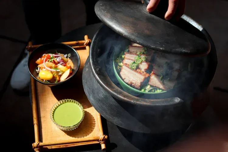 Black pot with soup beside two small bowls, including green sauce, on a woven tray.