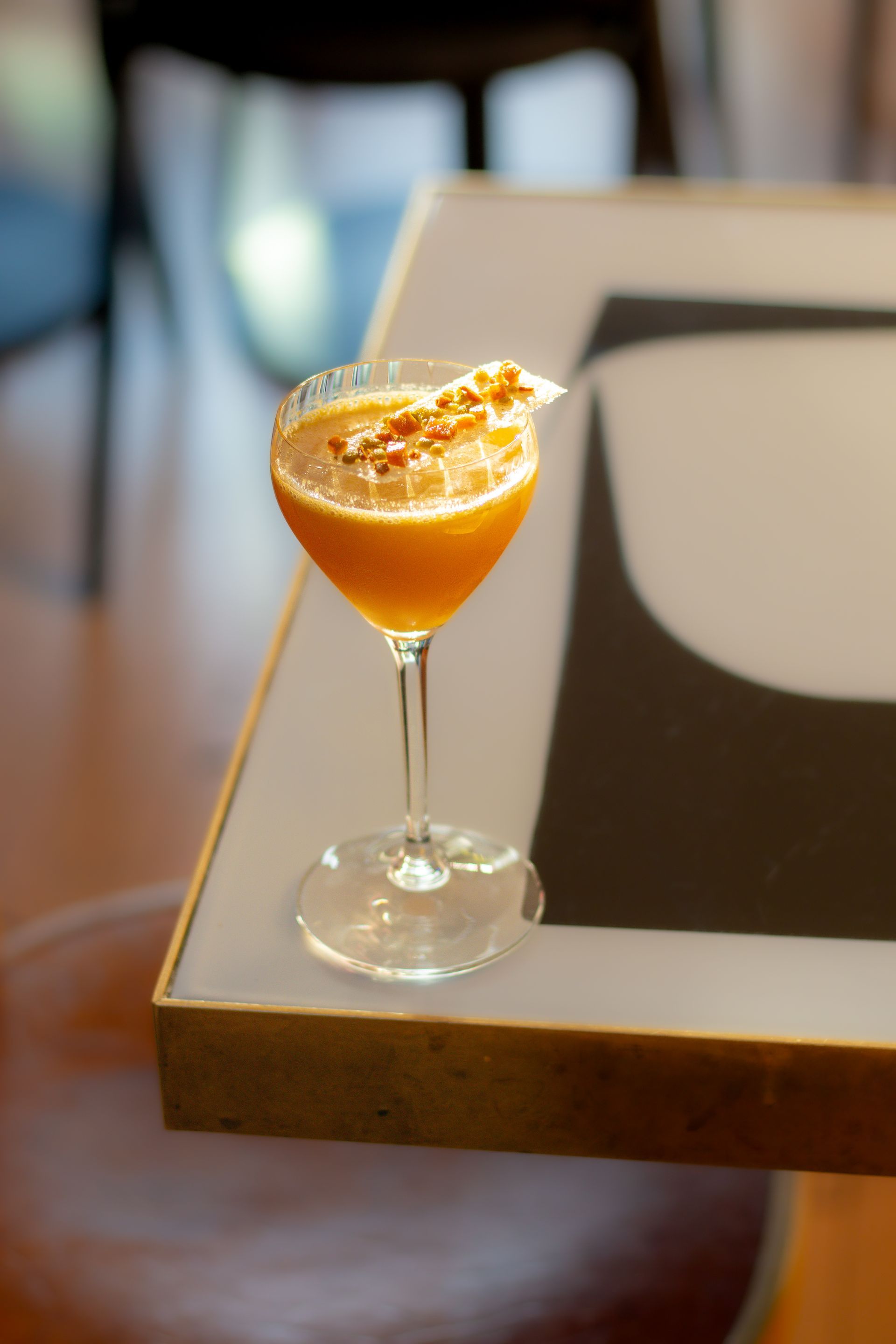 Orange cocktail in a stemmed glass with garnish on a table, blurred indoor background
