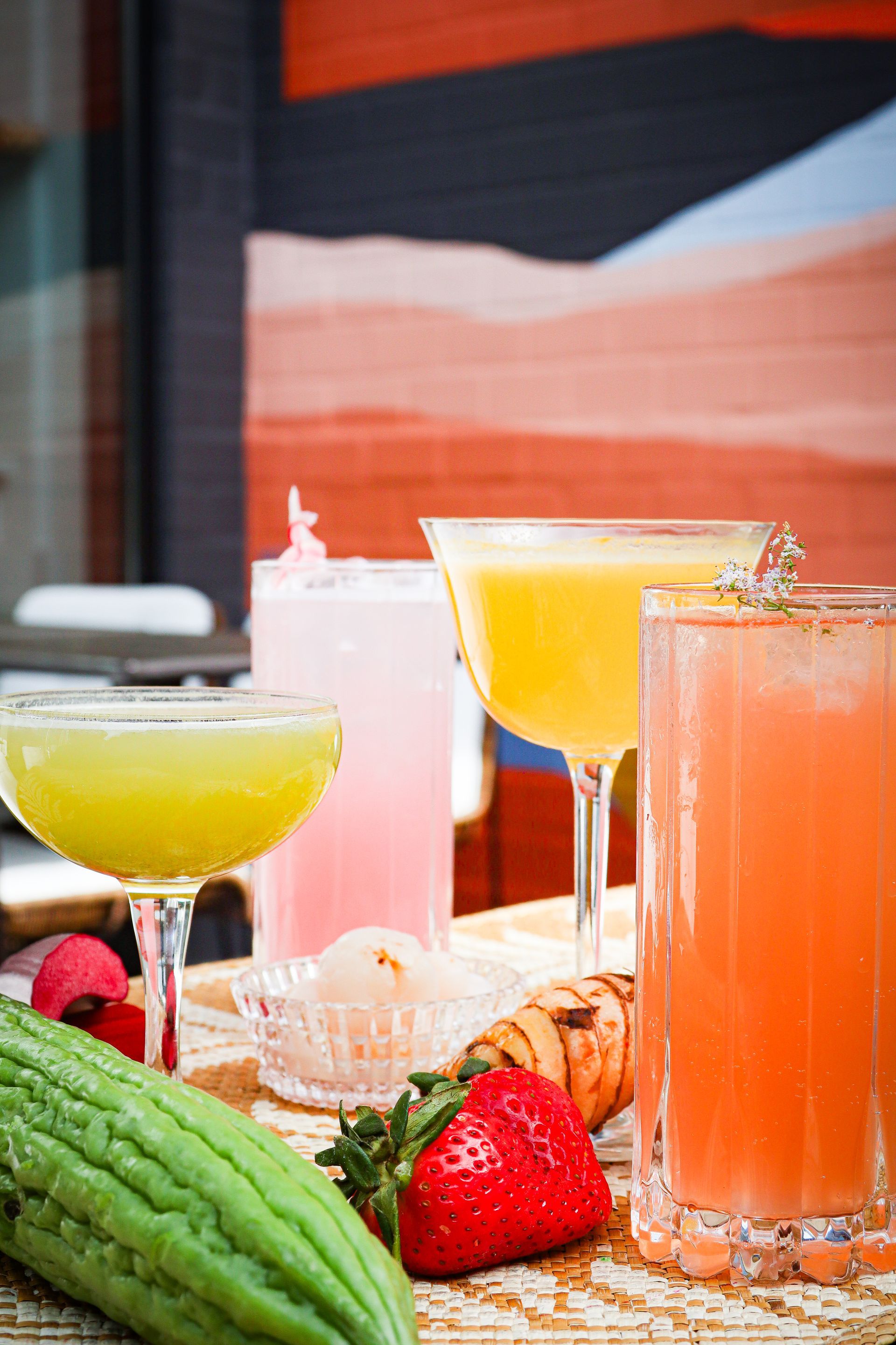 Assorted colorful cocktails and fruit on a table, including strawberries and a bitter melon, against a warm backdrop