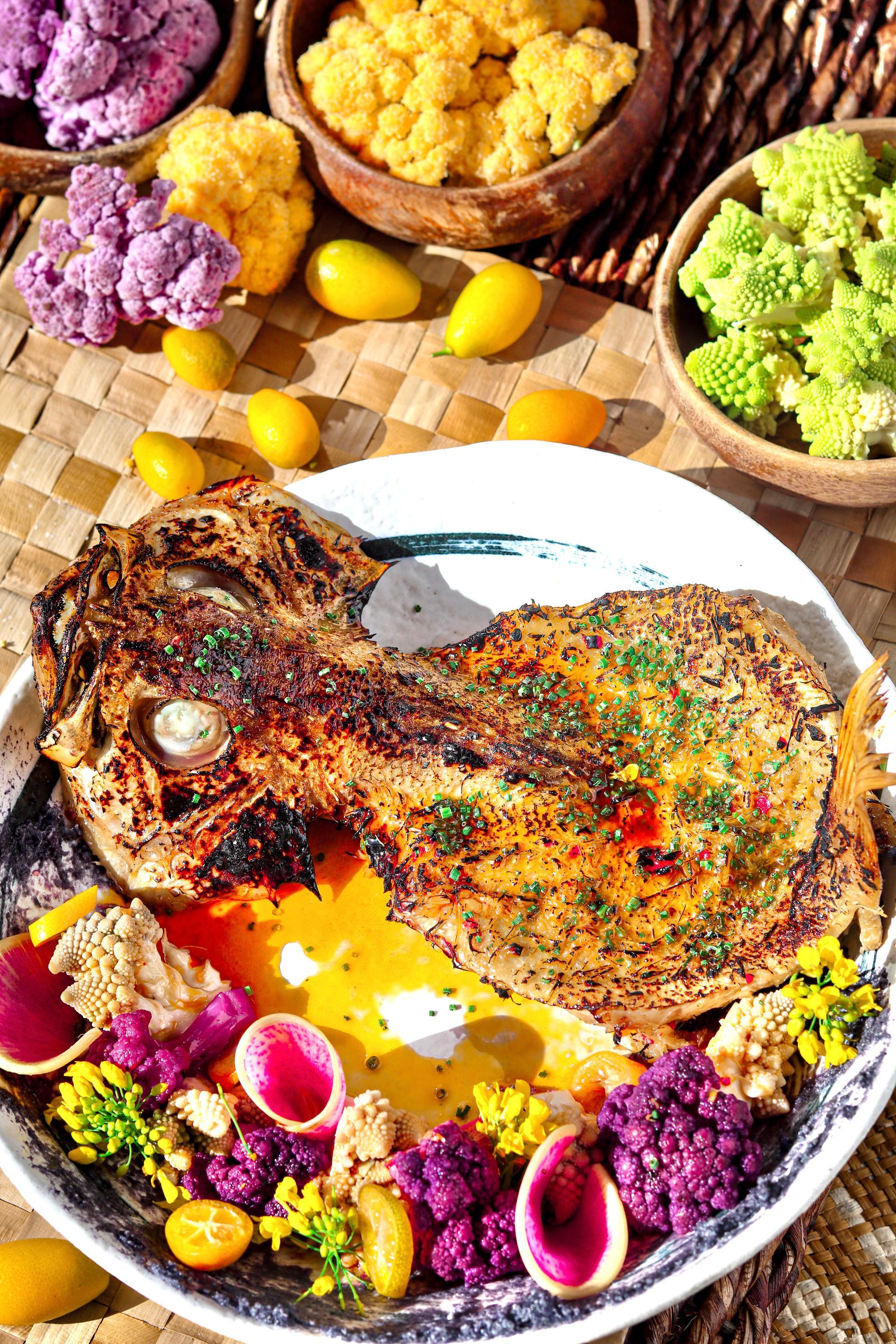 Grilled fish fillet on a white plate with colorful flower petals and small yellow tomatoes, surrounded by bowls of spices