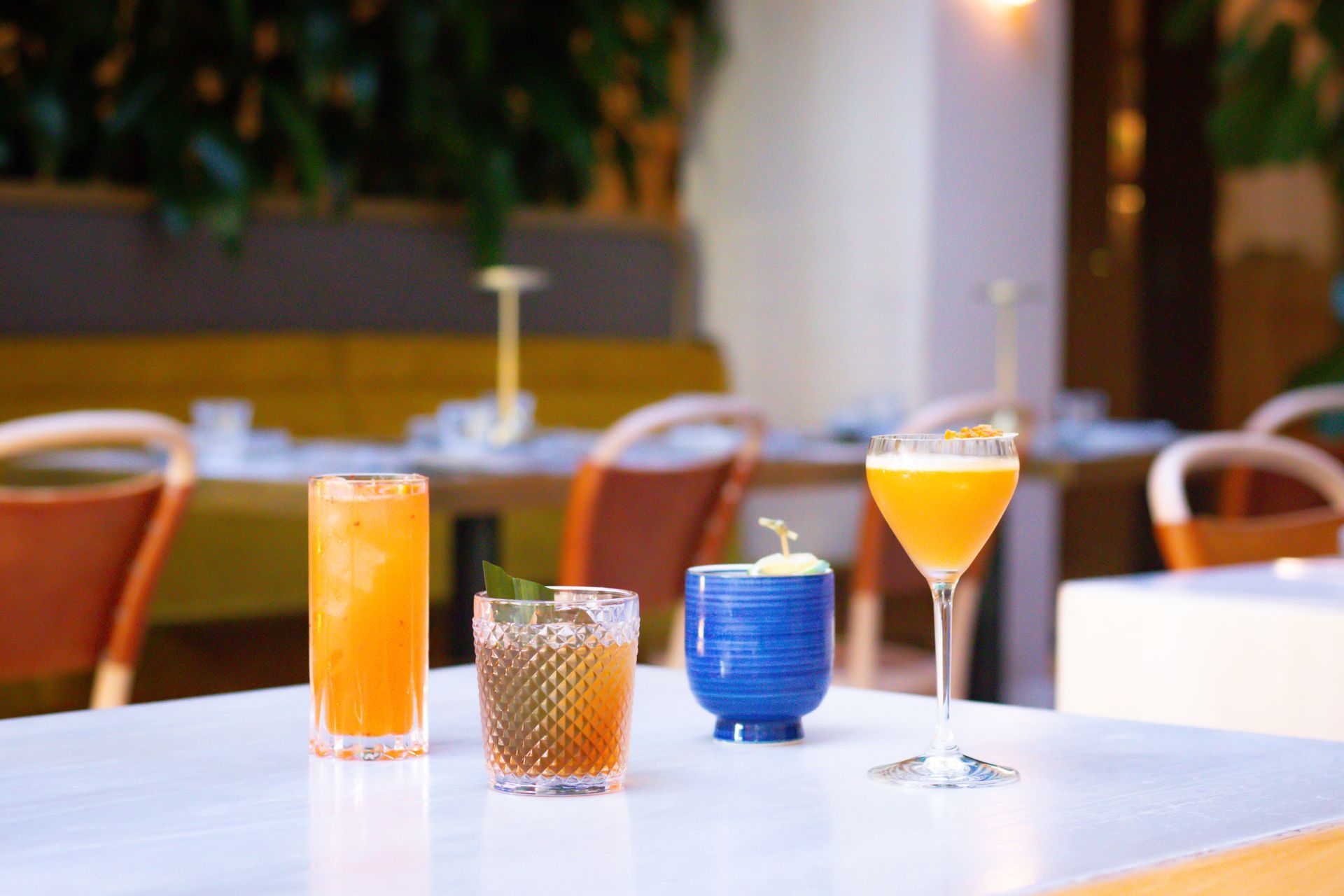 Four colorful cocktails on a white table in a softly lit bar or café.