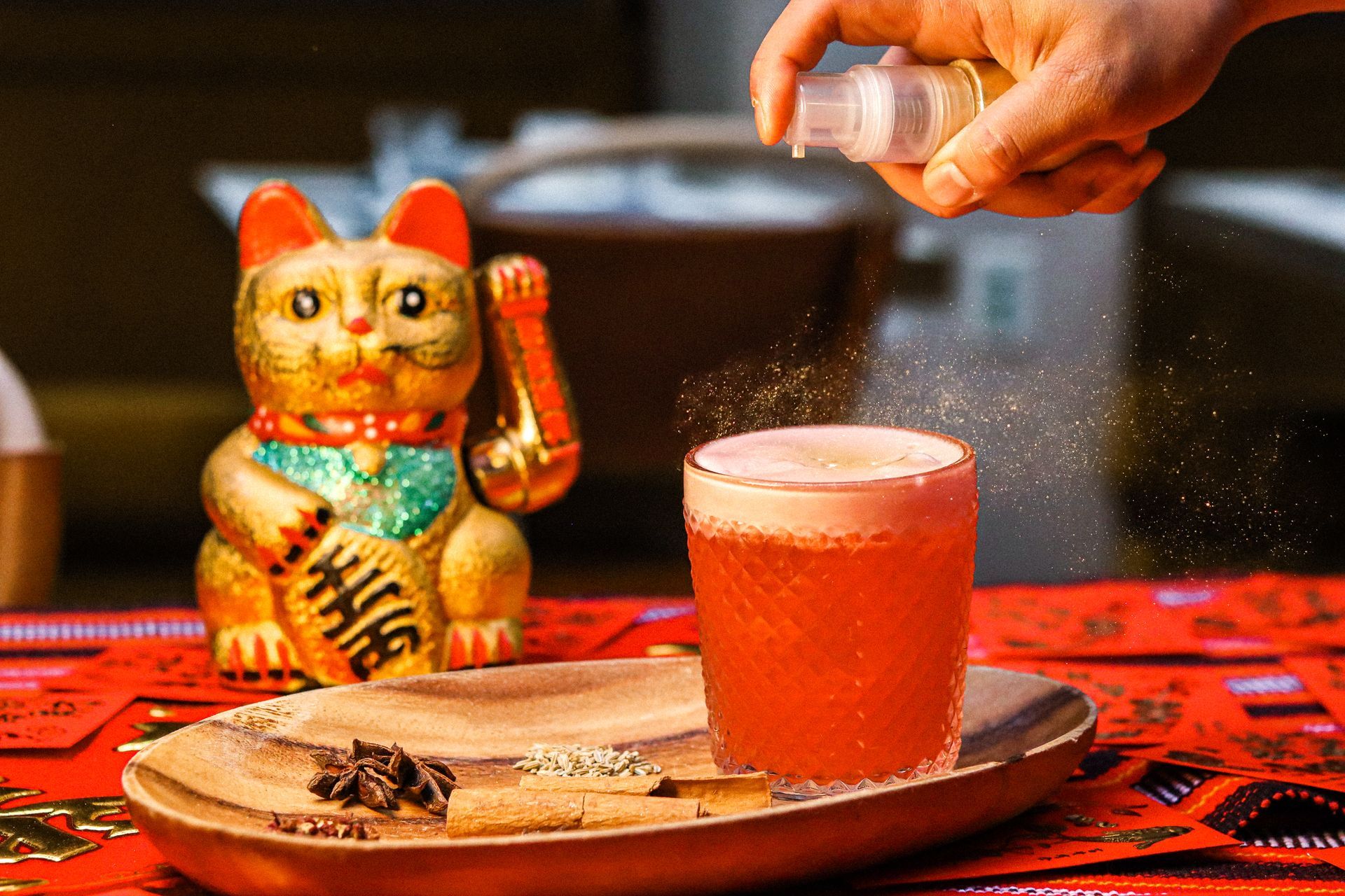 Cat figurine on a red table beside a frothy drink, with a hand sprinkling seasoning overhead.