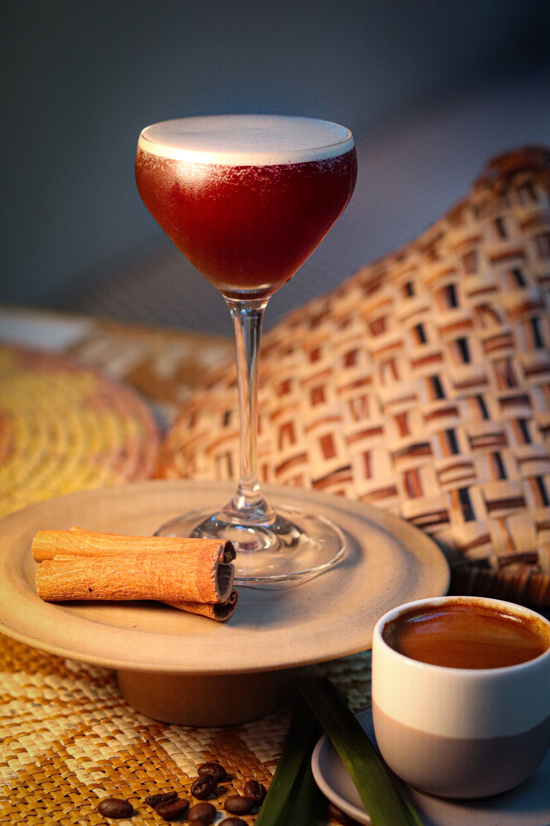 Cocktail in a coupe glass beside a cinnamon stick and espresso on a woven placemat