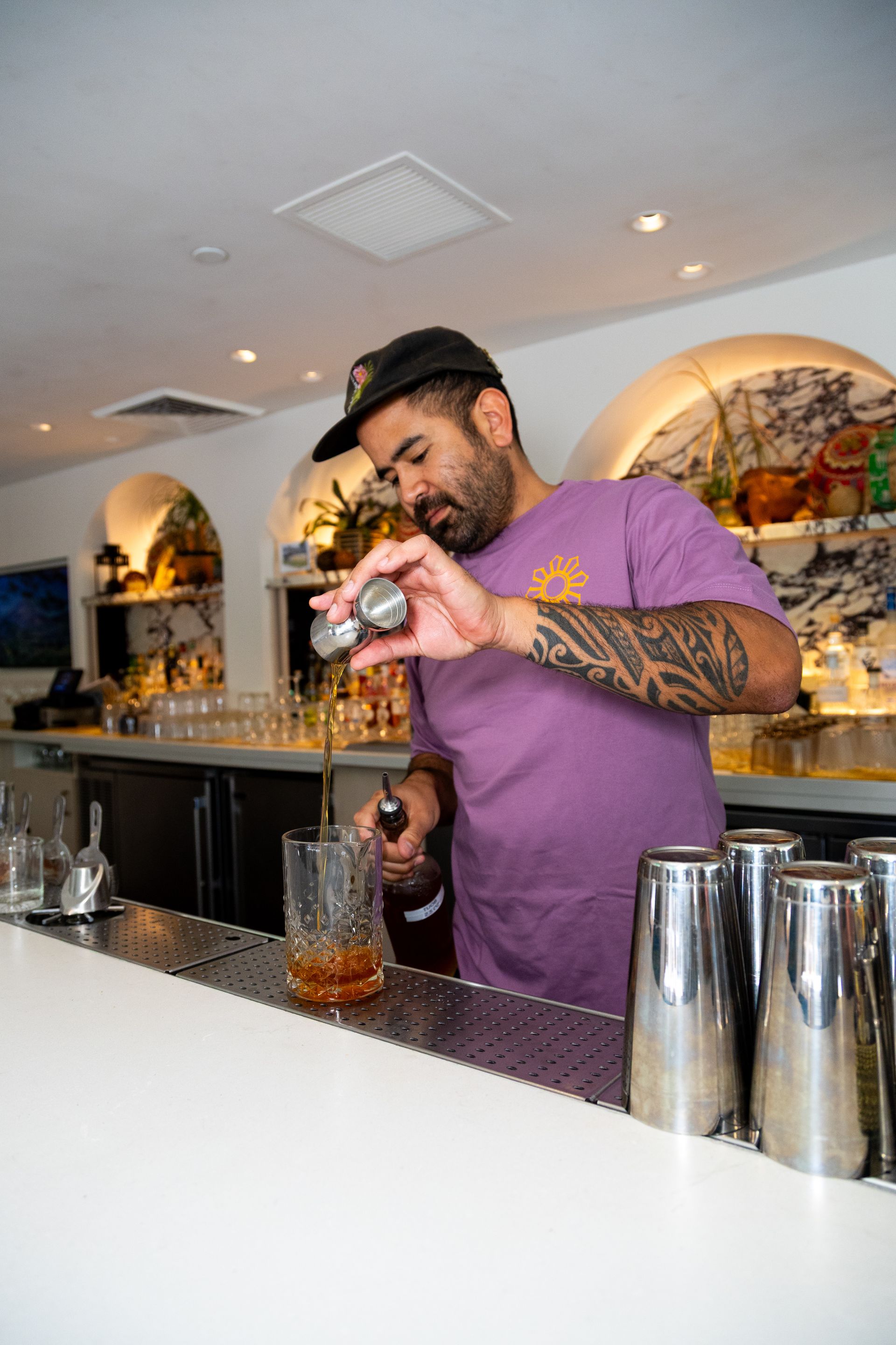 Bartender in a purple shirt shaking a drink behind a bar with cocktail shakers and glassware.