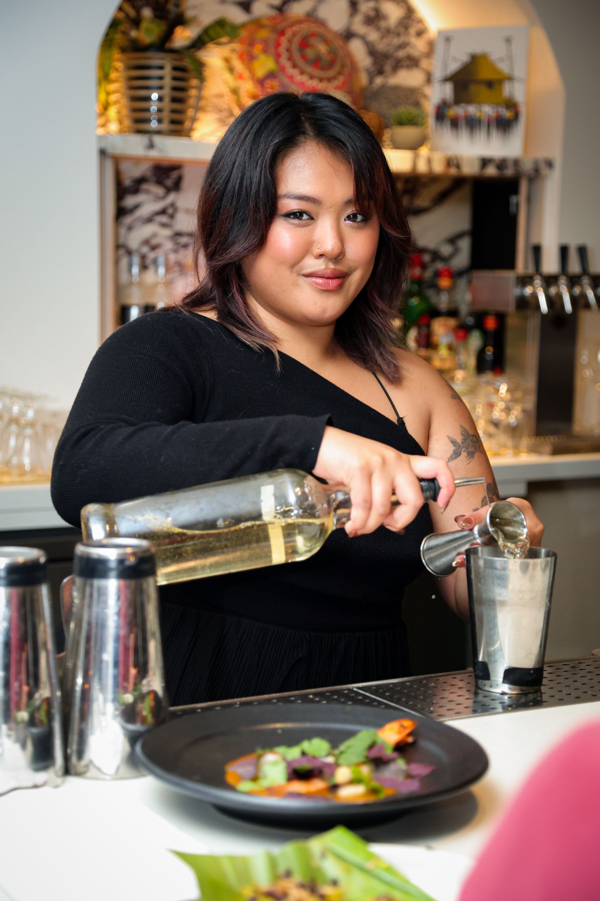 Bartender pouring a clear drink into a cocktail shaker at a bar counter