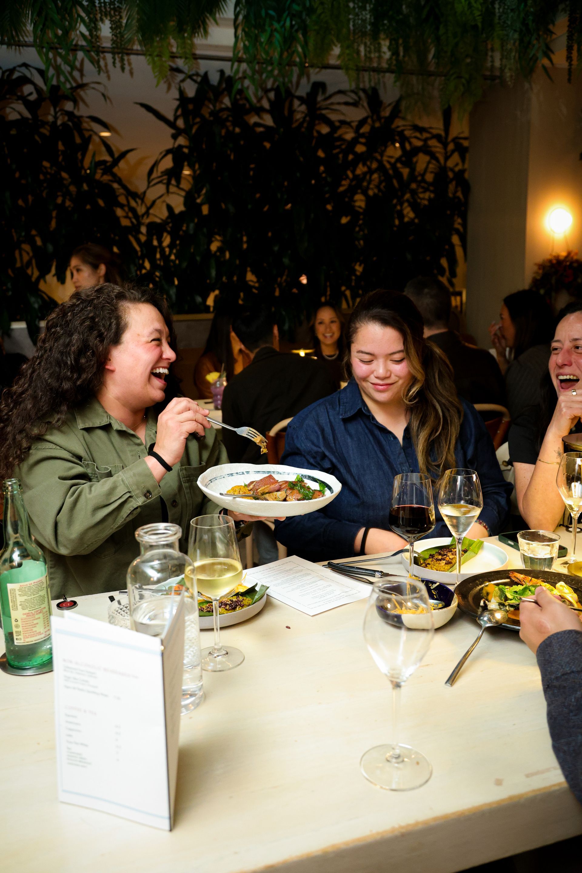 Friends laughing over dinner at a dimly lit restaurant table with wine glasses and dishes