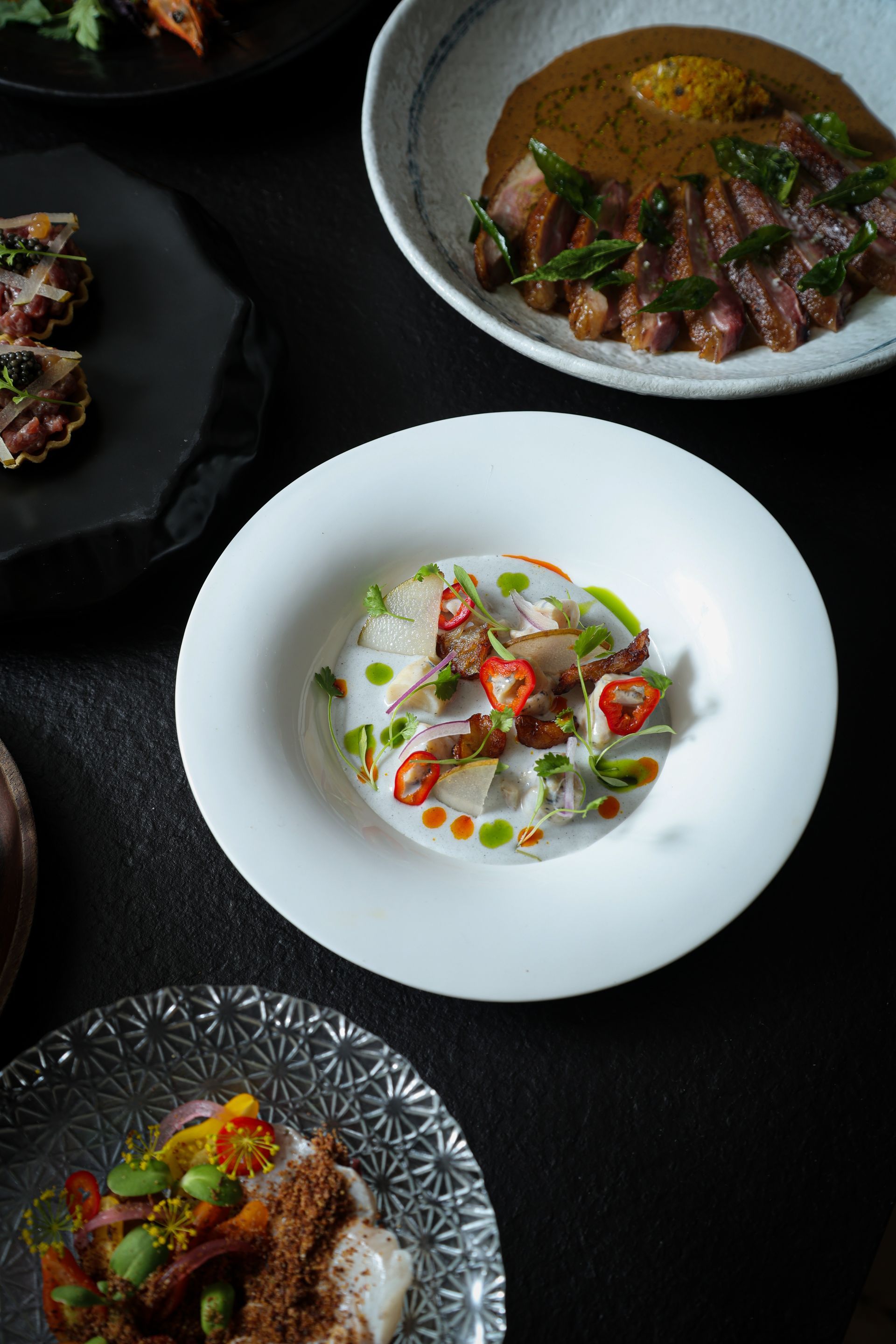 Plated gourmet dishes on dark tableware, including a white bowl with fish, herbs, and cherry tomatoes