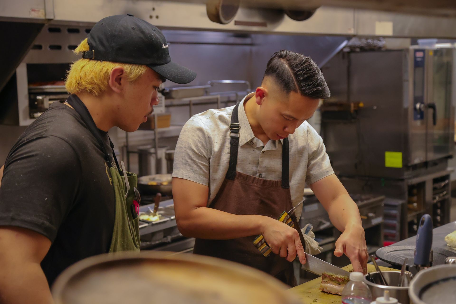 Two chefs preparing food together in a commercial kitchen, one chopping ingredients beside a stovetop.