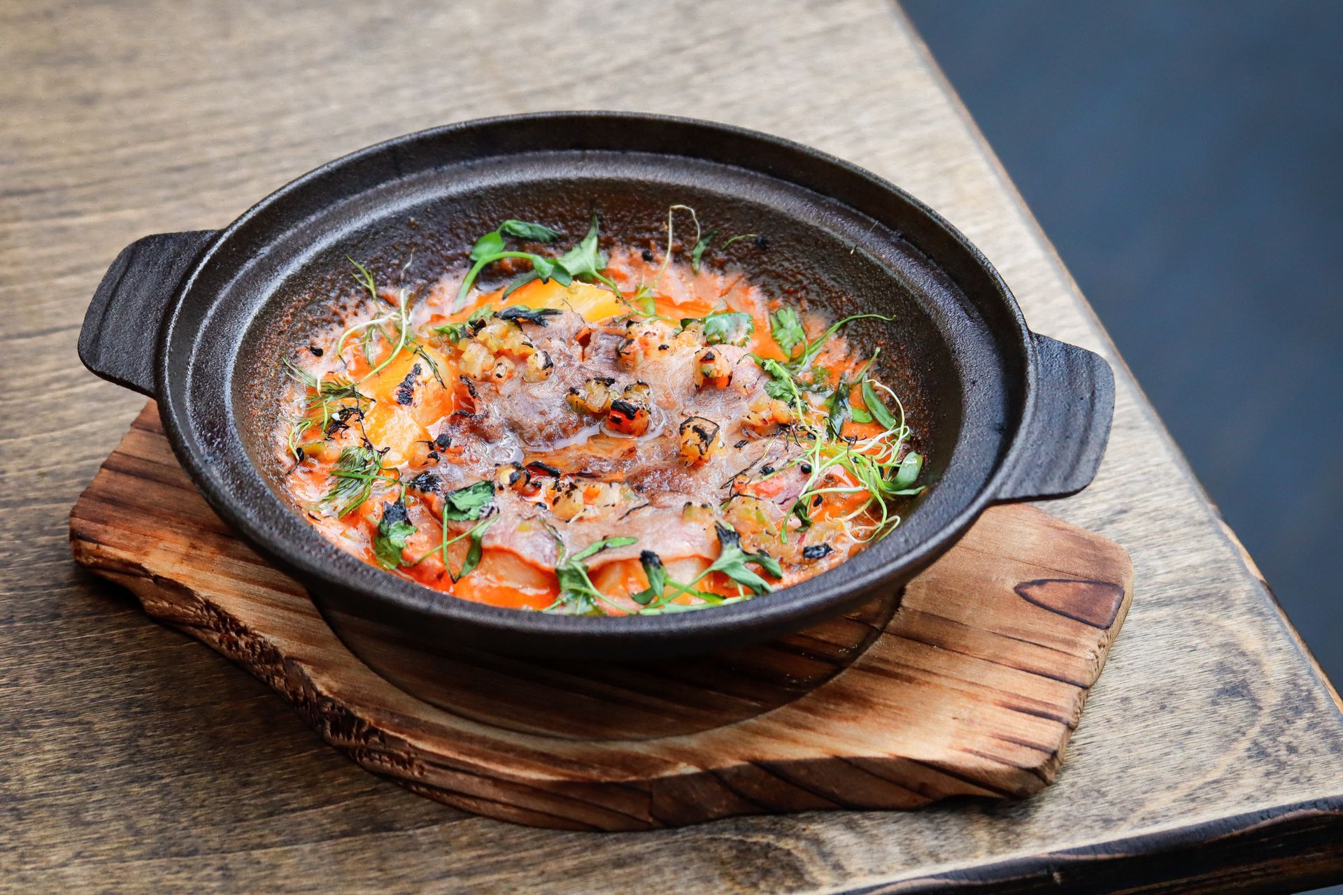 Black clay pot of stew garnished with herbs, sitting on a wooden board and stone table.