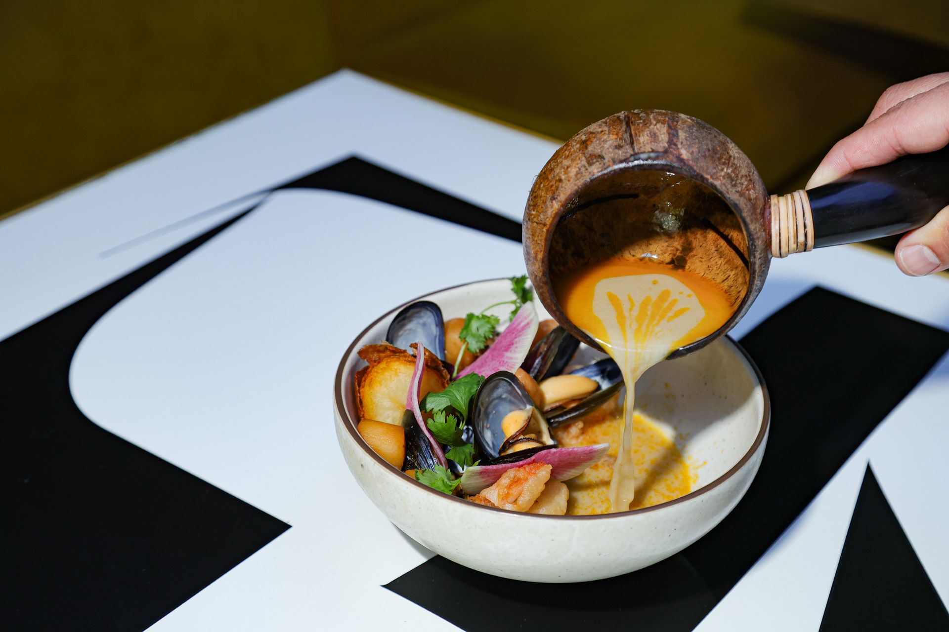 Hand pouring broth into a bowl of mussels and vegetables on a black-and-white table