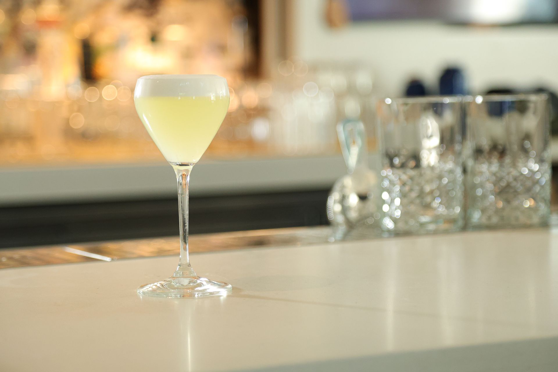 Pale yellow cocktail in a martini glass on a bar counter with blurred glasses in the background