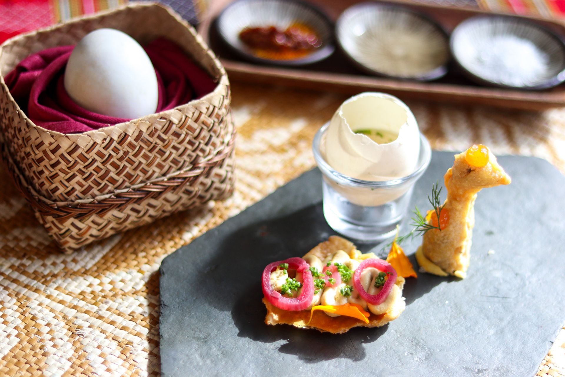 Restaurant table with a woven basket, small appetizer on a black slate, and condiments in the background