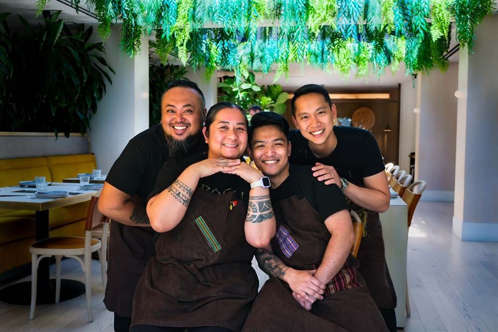 Four people in black shirts smiling and hugging at a decorated indoor table with hanging greenery.