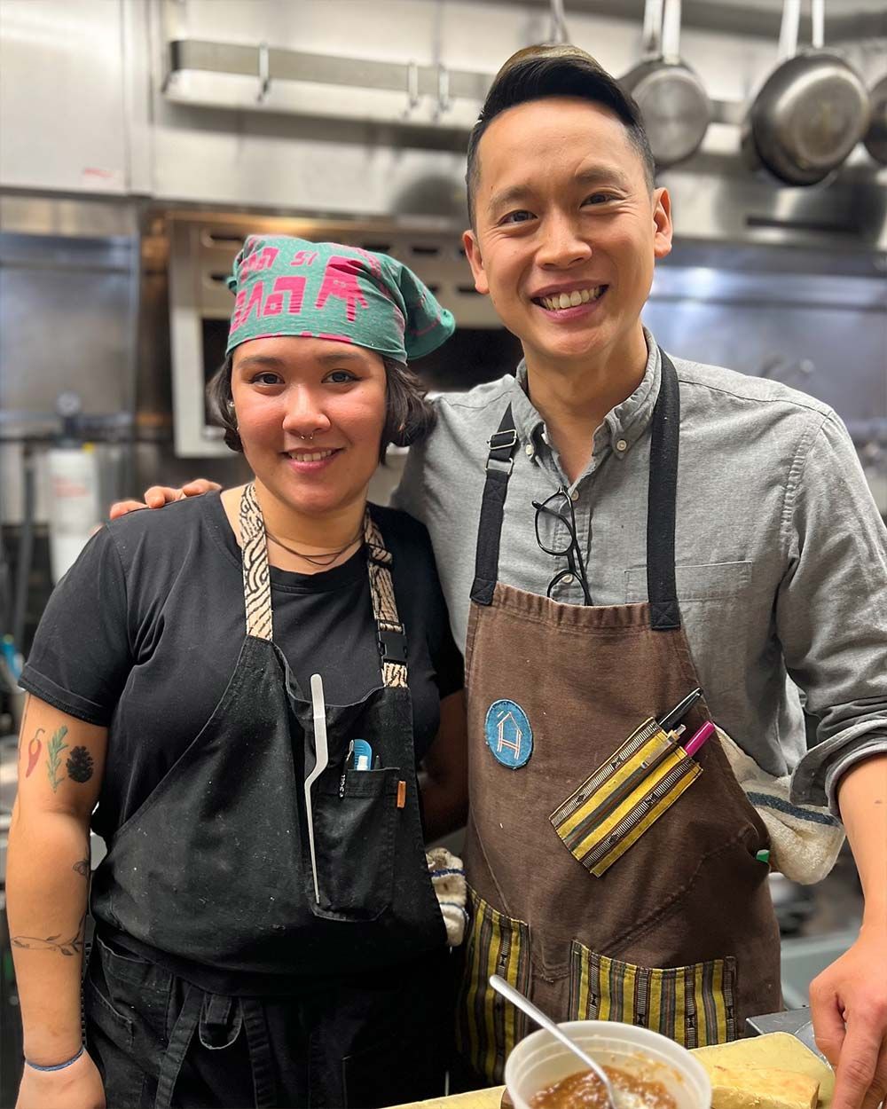 Two smiling chefs in aprons posing together in a kitchen, one holding a bowl with utensils.