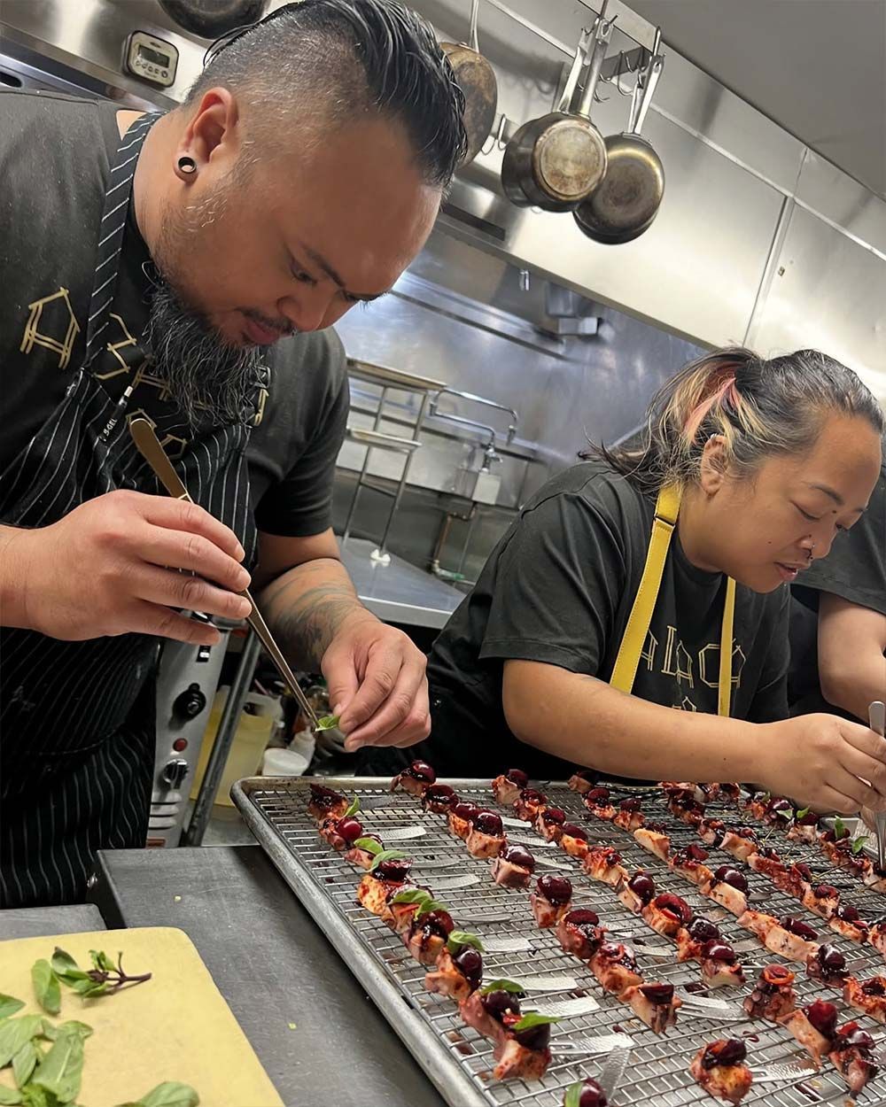 Two chefs plating rows of bite-sized appetizers in a busy stainless-steel kitchen.