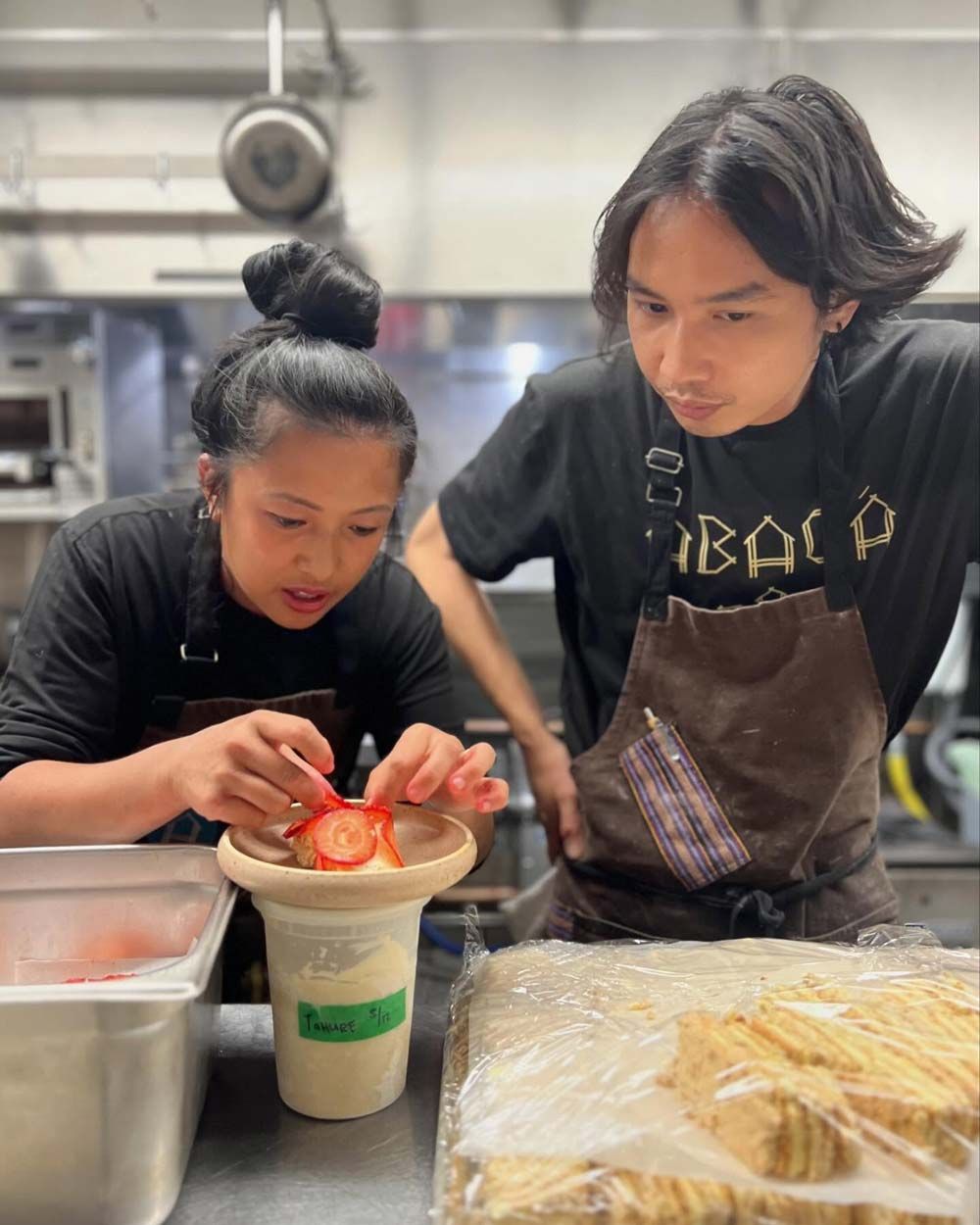 Two kitchen staff preparing food, slicing strawberries and dough at a stainless-steel counter.