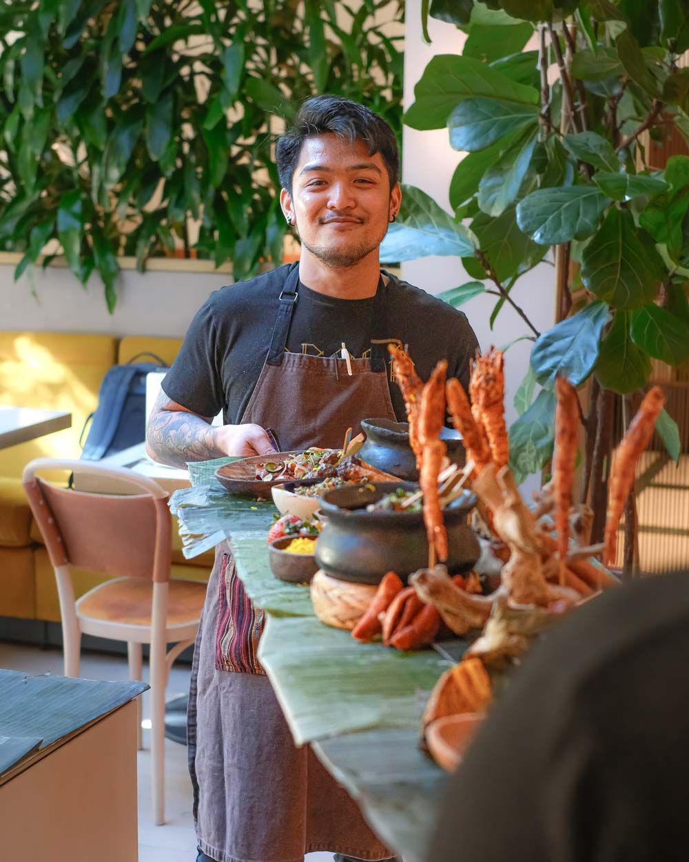 A smiling person stands behind a seafood platter at a restaurant table with leafy decor.