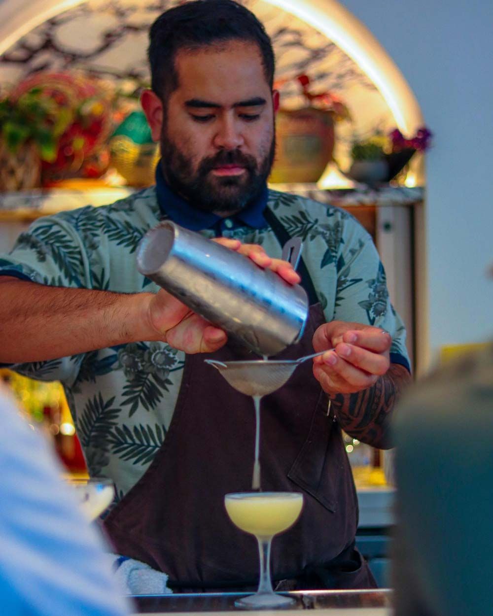 Bartender straining a cocktail into a coupe glass behind a bar, with a yellow drink below.