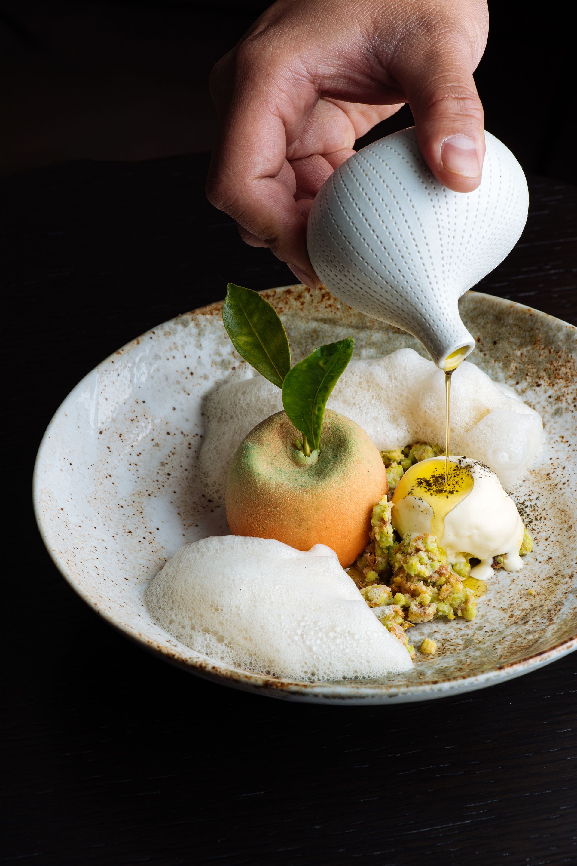 Hand pouring white sauce over a plated fruit dessert with apple, mint, and granola