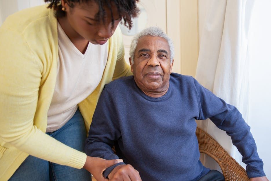 An elderly man is sitting in a chair while a woman holds his hand.