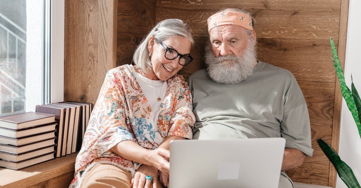 An elderly couple is sitting on a window sill looking at a laptop computer.