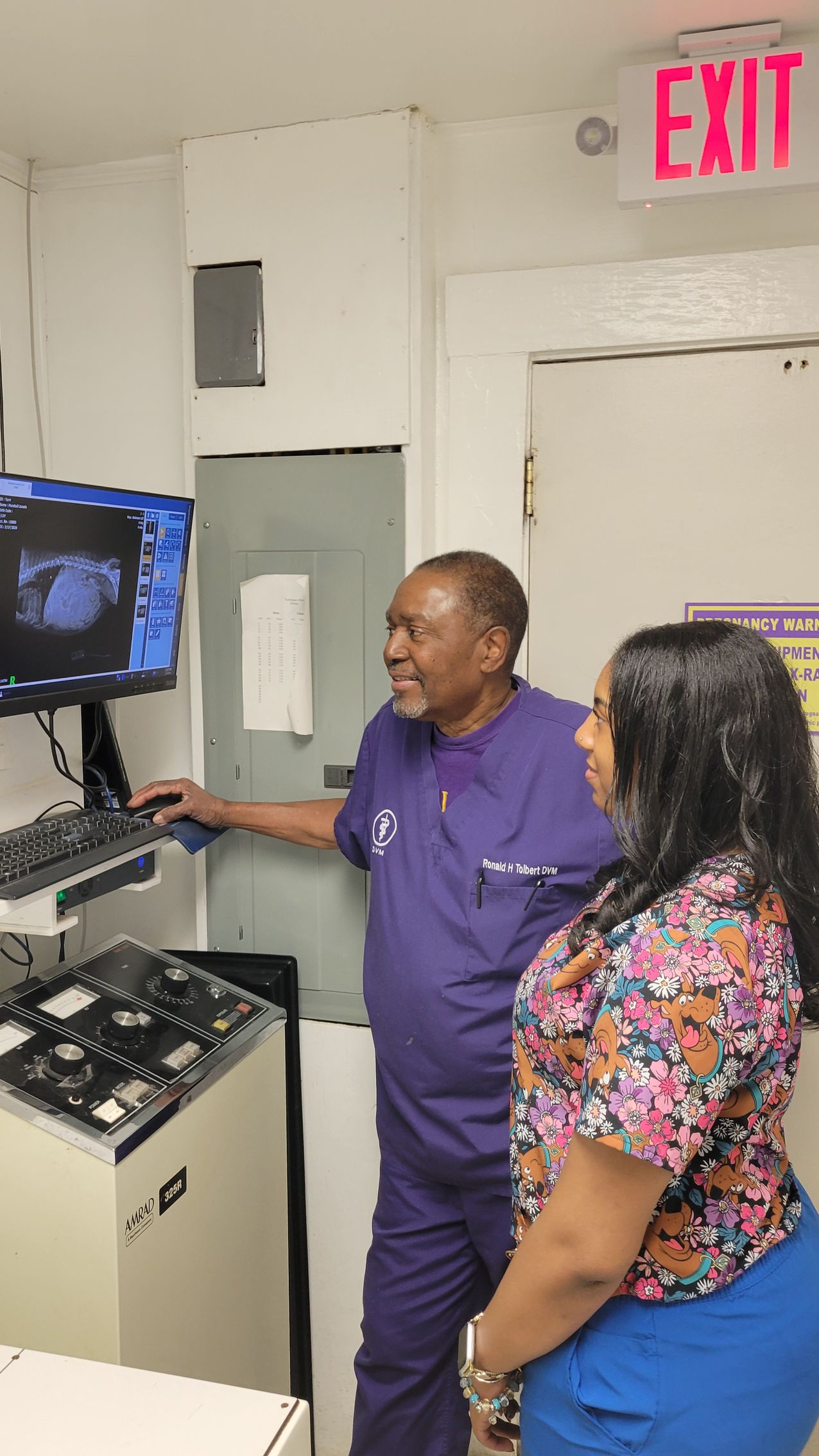 Man in purple scrubs points at a medical screen, woman looks on. Inside room, Exit sign.