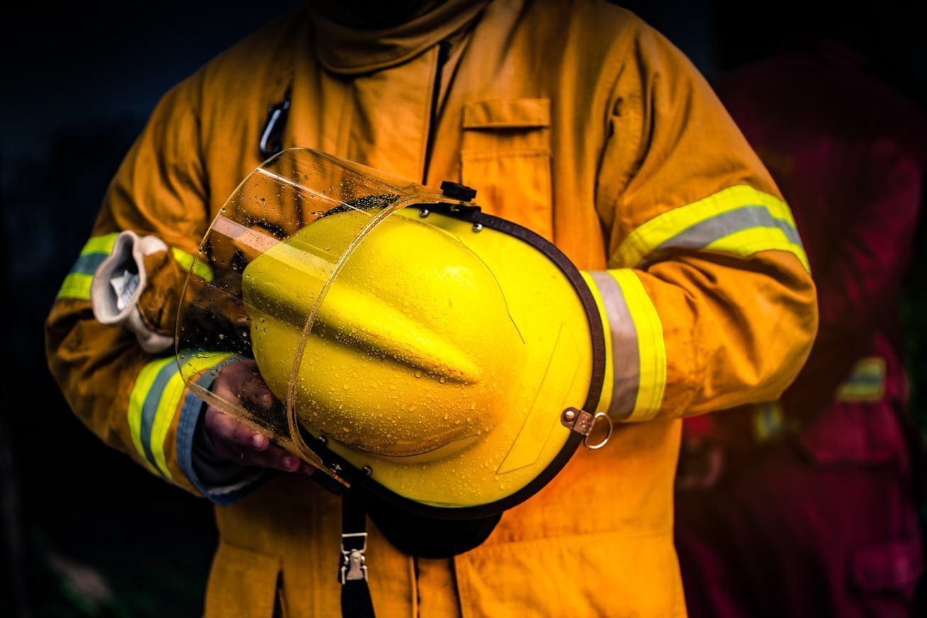 Firefighter in Yellow Coat Holding a Yellow Hard Hat in a Dark Setting — Safe Industries Group in Woodrising, NSW
