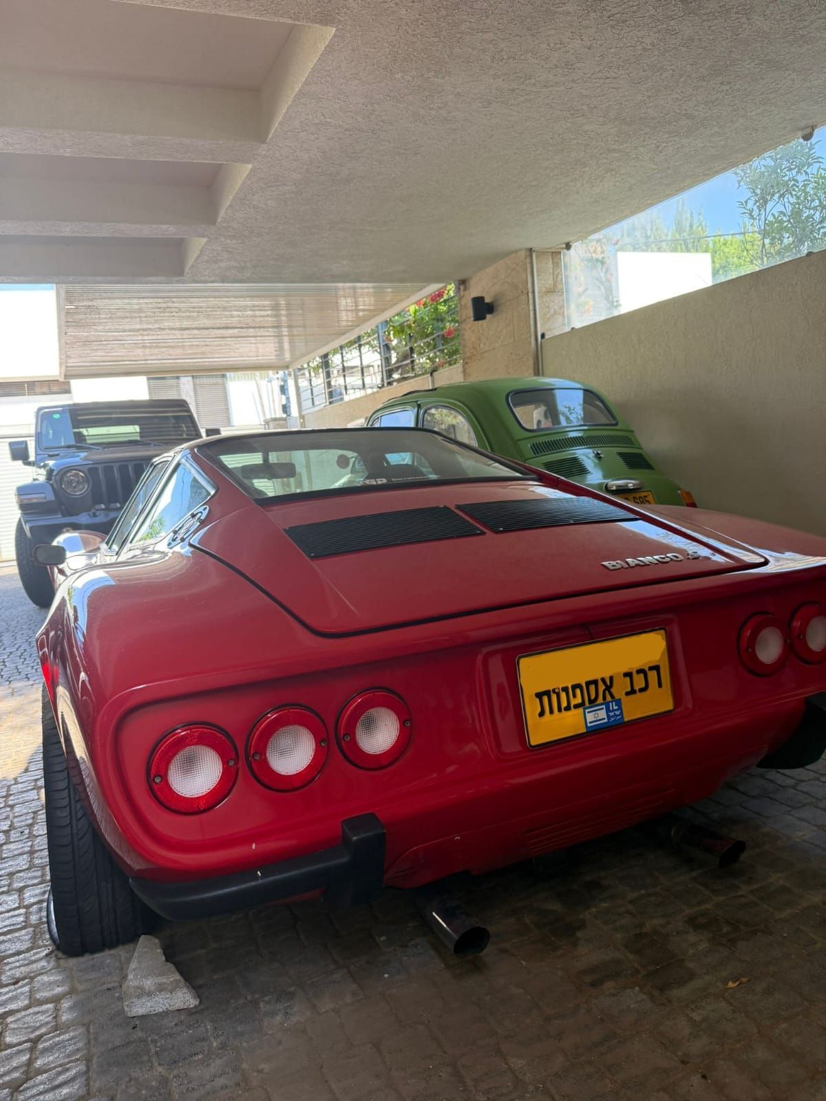 מכונית ספורט קלאסית אדומה חונה מתחת לשביל גישה מקורה, מבט אחורי עם לוחית רישוי צהובה.