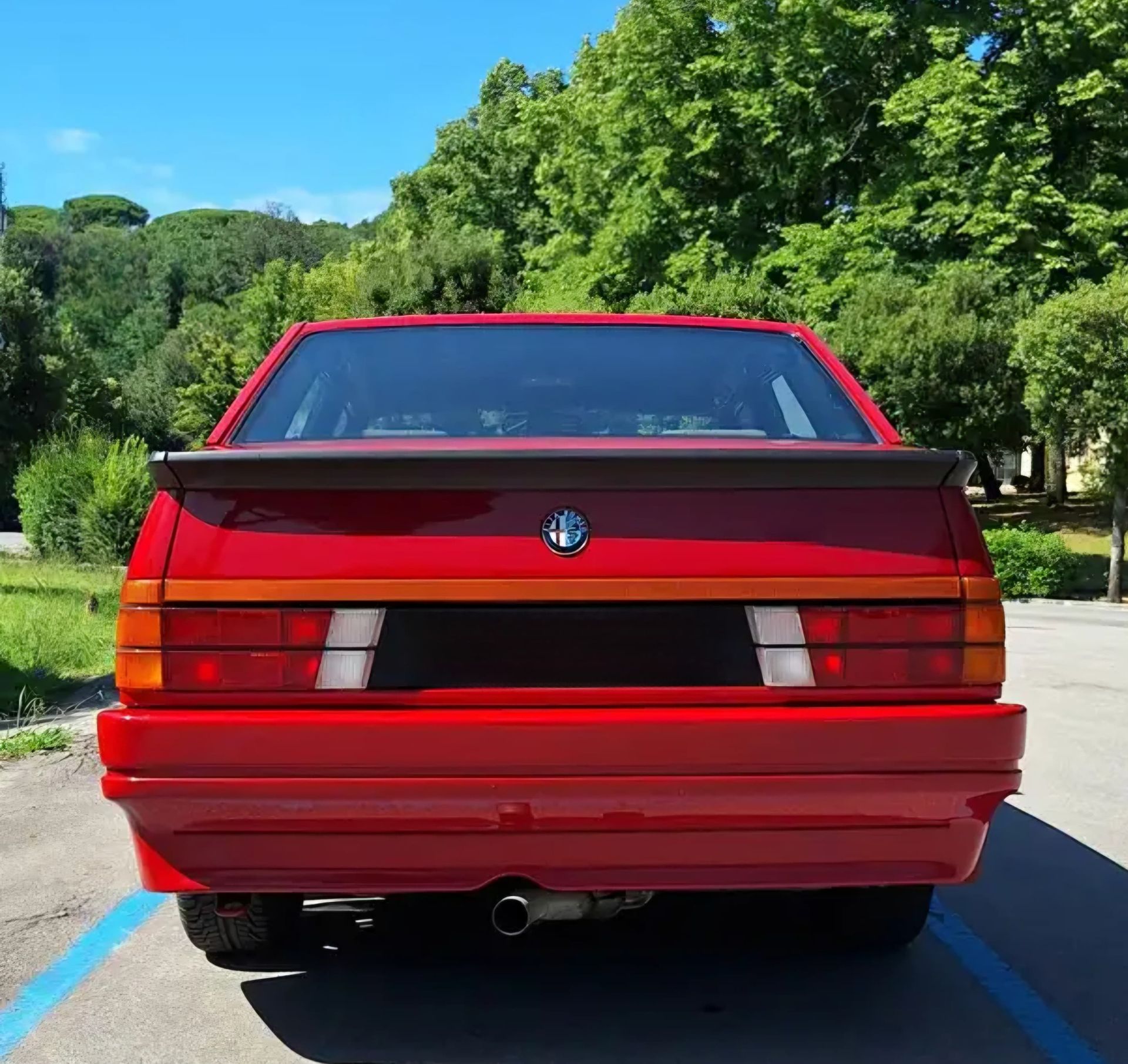 מכונית קופה אדומה של BMW שנראית מאחור על כביש שטוף שמש עם עצים ברקע
