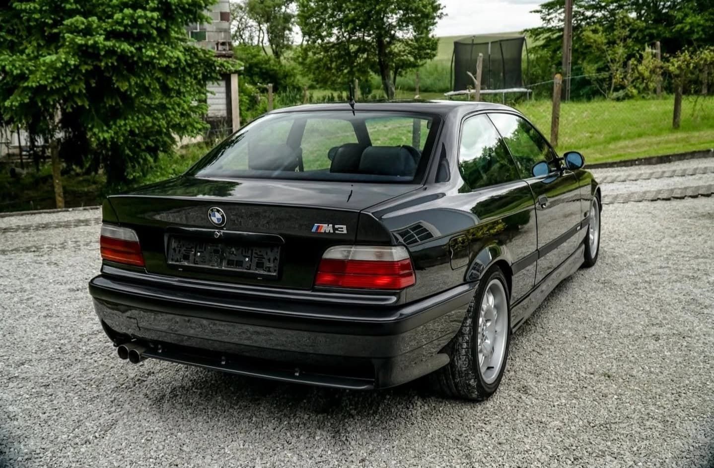 מכונית סדאן שחורה של BMW חונה על שביל גישה חצץ עם עצים ובית ברקע