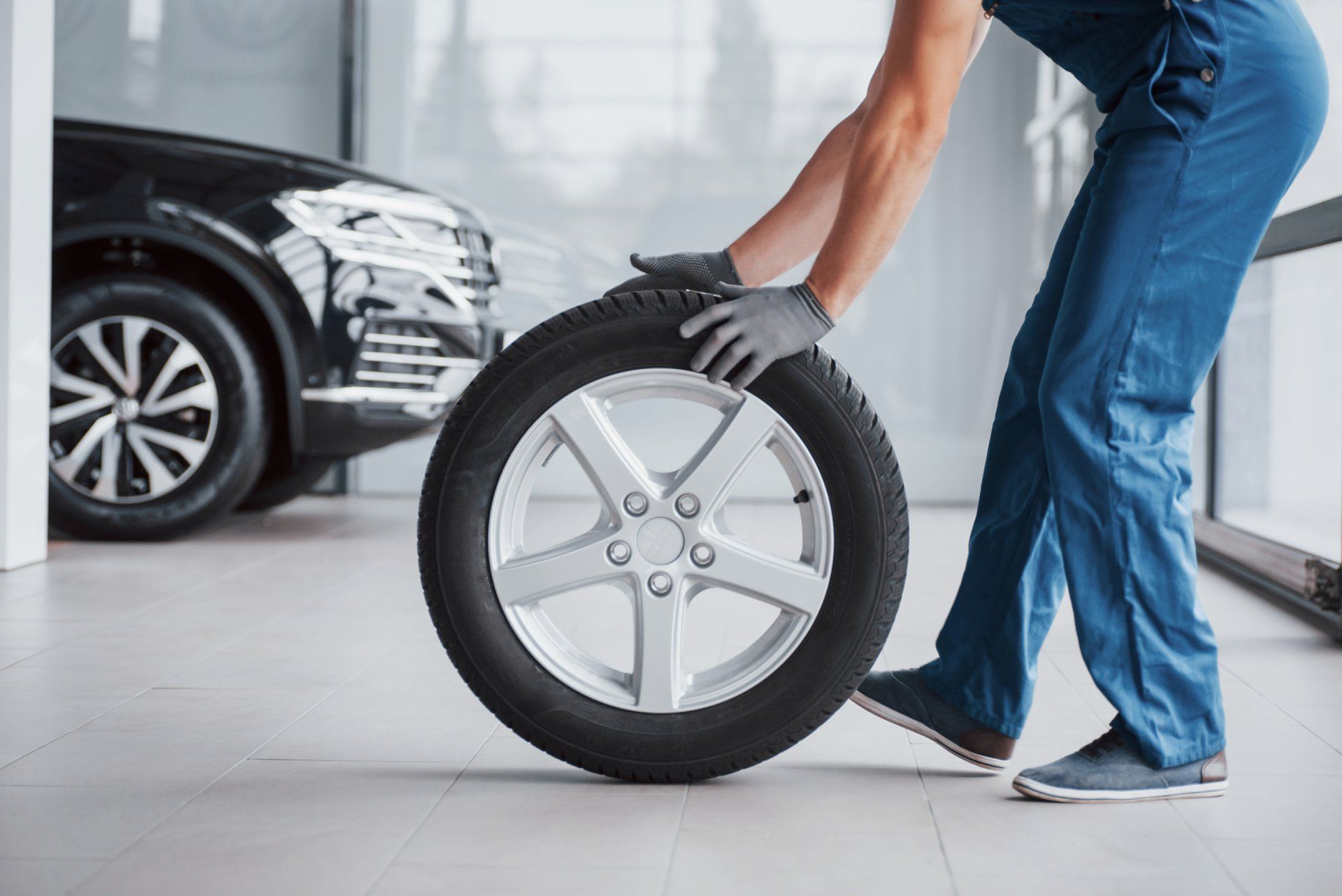 Un mécanicien en combinaison bleue fait rouler un pneu à jante argentée dans un atelier de réparation automobile.