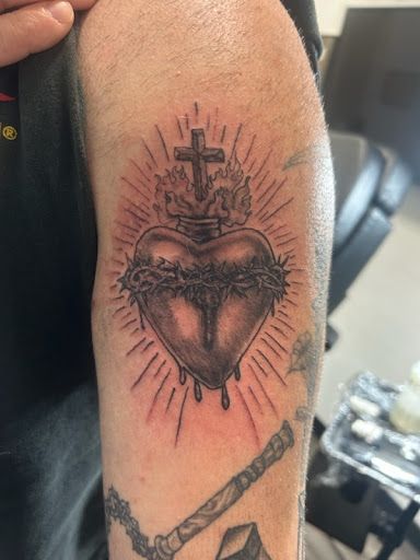Upper arm tattoo of a heart with a cross and radiant lines, with a chain-like tattoo below