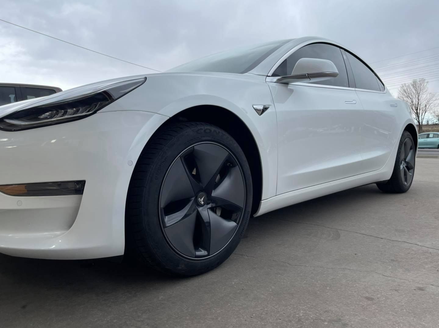 A white Tesla Model 3 parked on a concrete surface under an overcast sky.