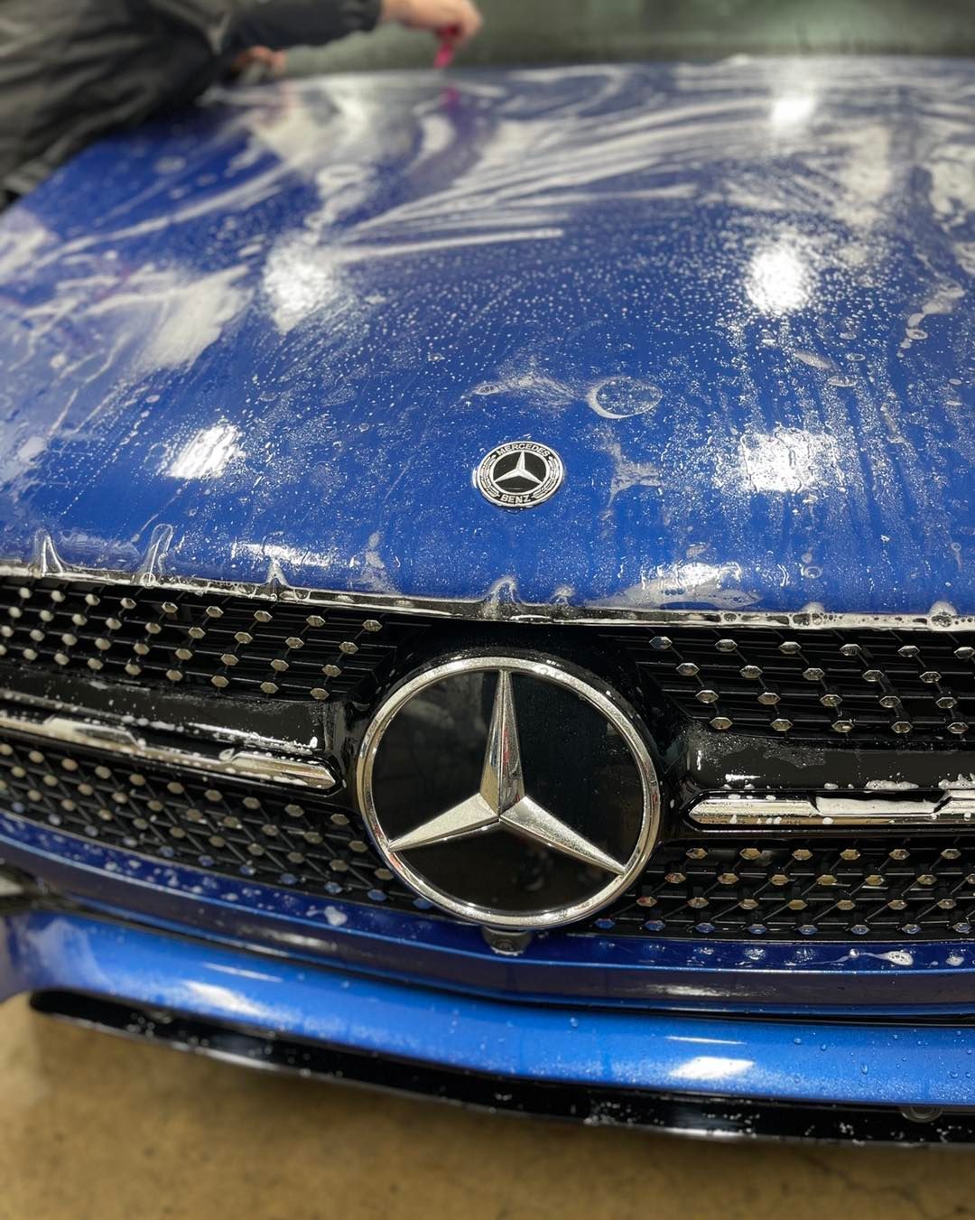 A person washes a blue Mercedes-Benz with soap and water, focusing on the hood and front grille emblem.