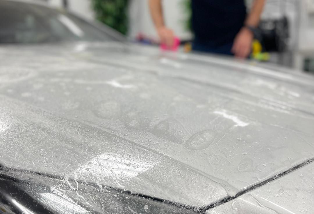 A person applies a white cleaning product to the hood of a silver car in a workshop setting.