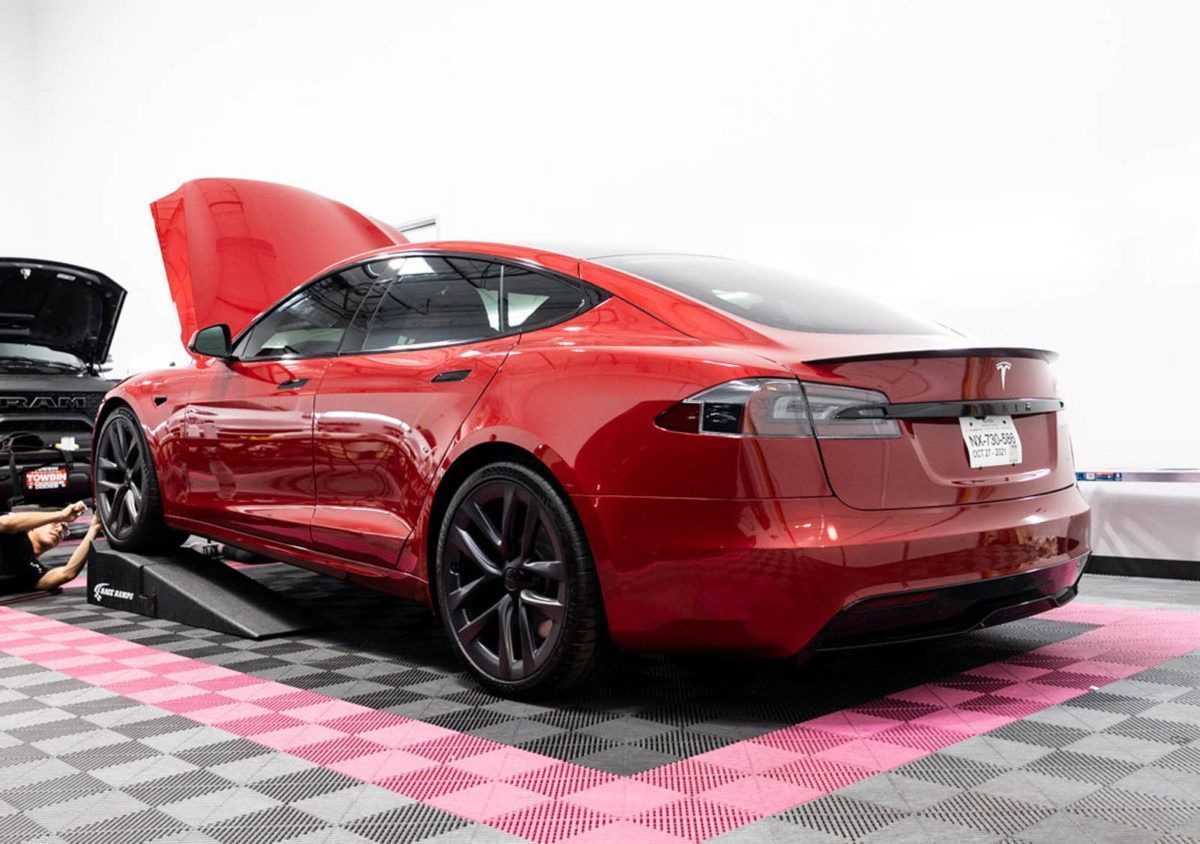 A red Tesla Model S with its front hood open, parked on ramps inside a garage with pink and gray checkered flooring.